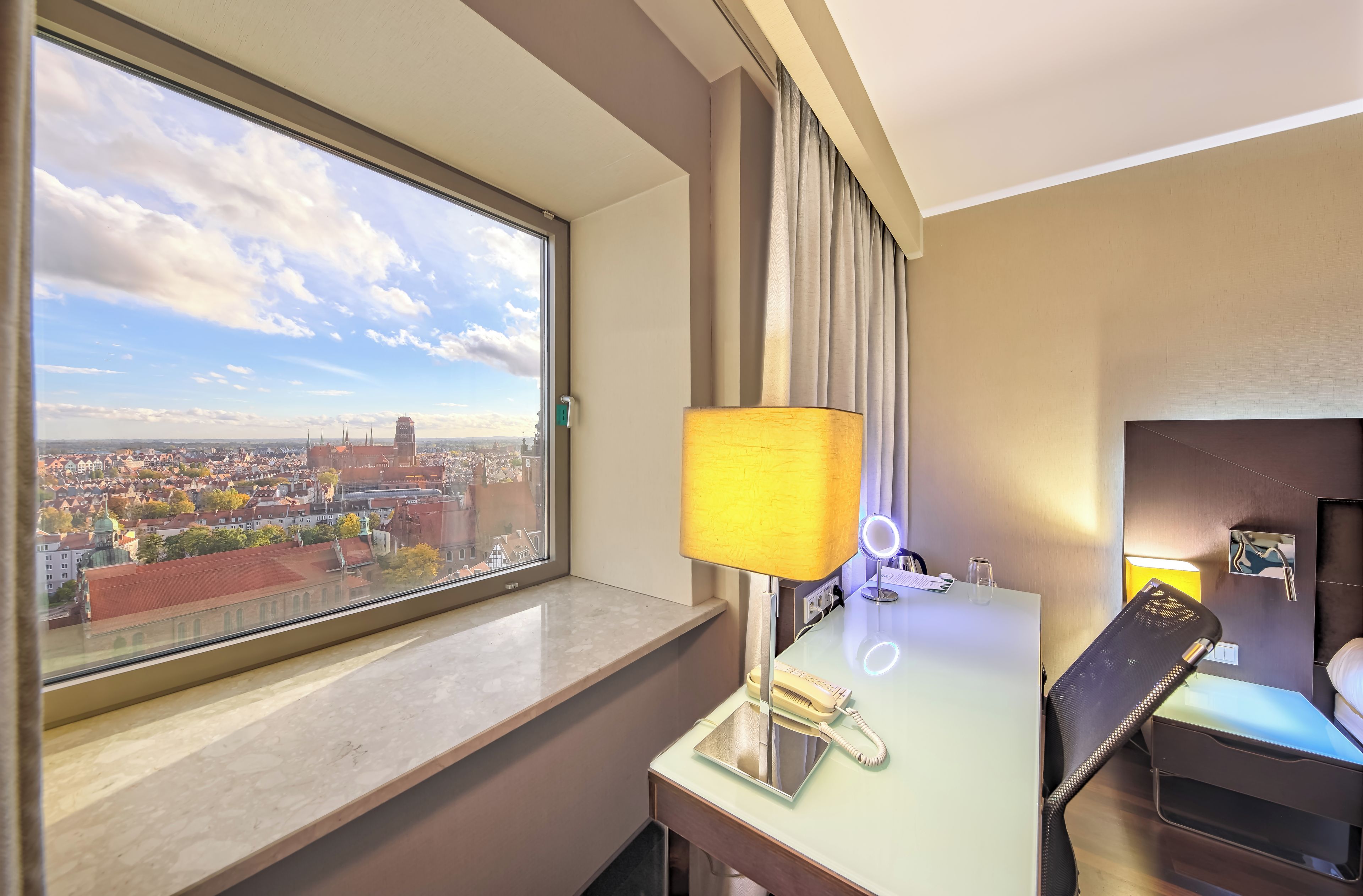 Una habitación de hotel con un ventanal grande, un escritorio con lámpara, un teléfono y una silla.
