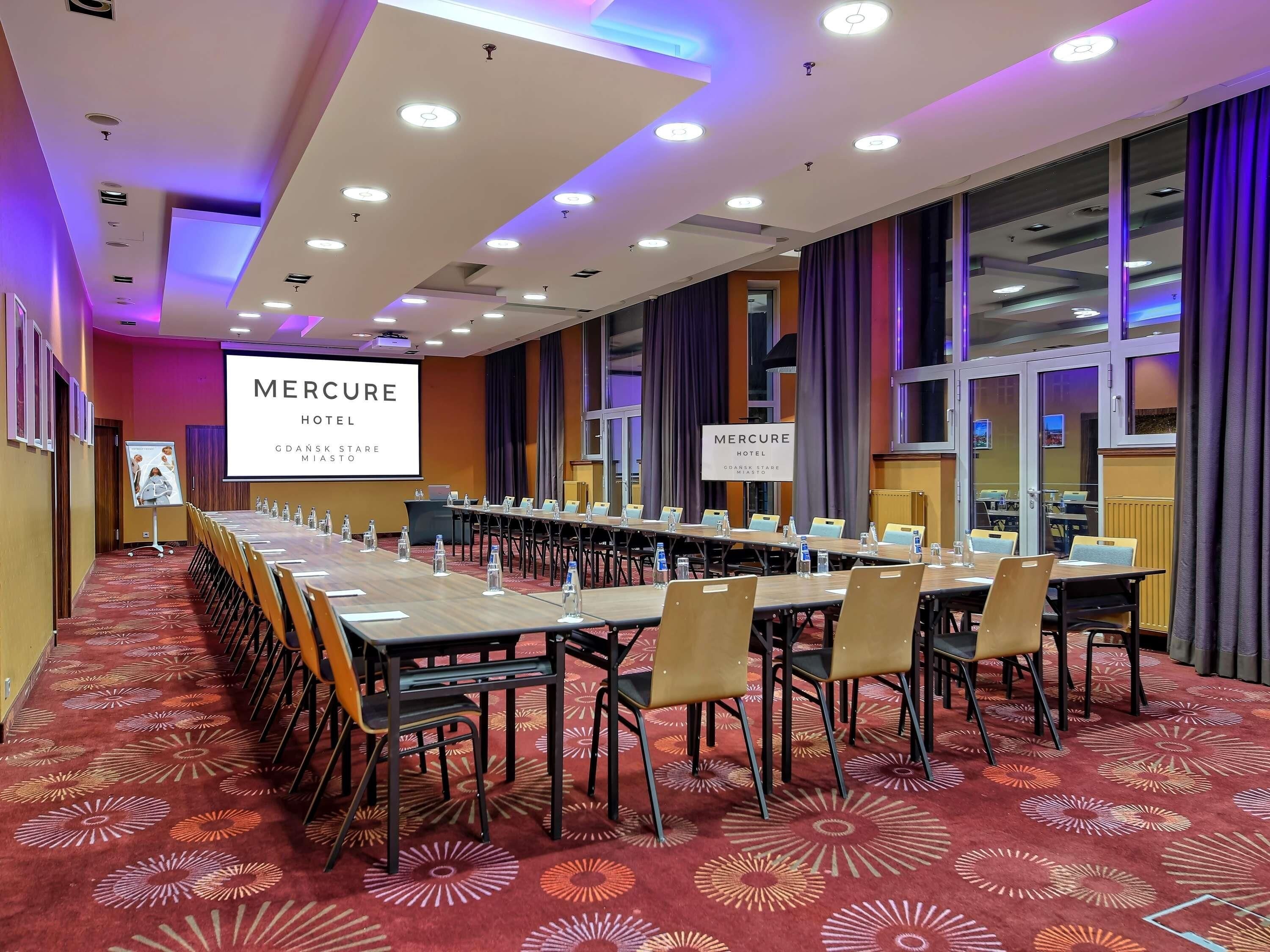 Ein Konferenzraum mit langem Tisch, Stühlen, Wasserflaschen und einem Bildschirm, der 'Mercure Hotel' anzeigt.