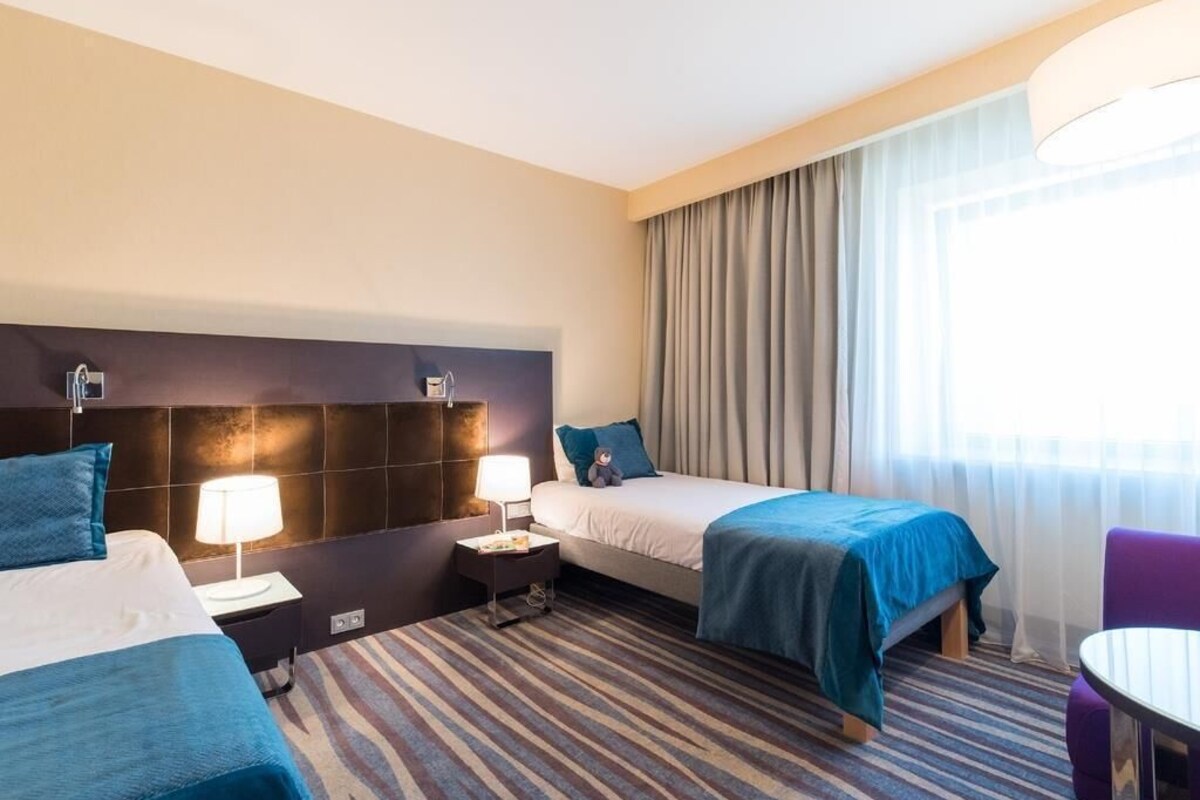 A hotel room with two beds, a desk, a chair, a lamp, and a window with curtains.