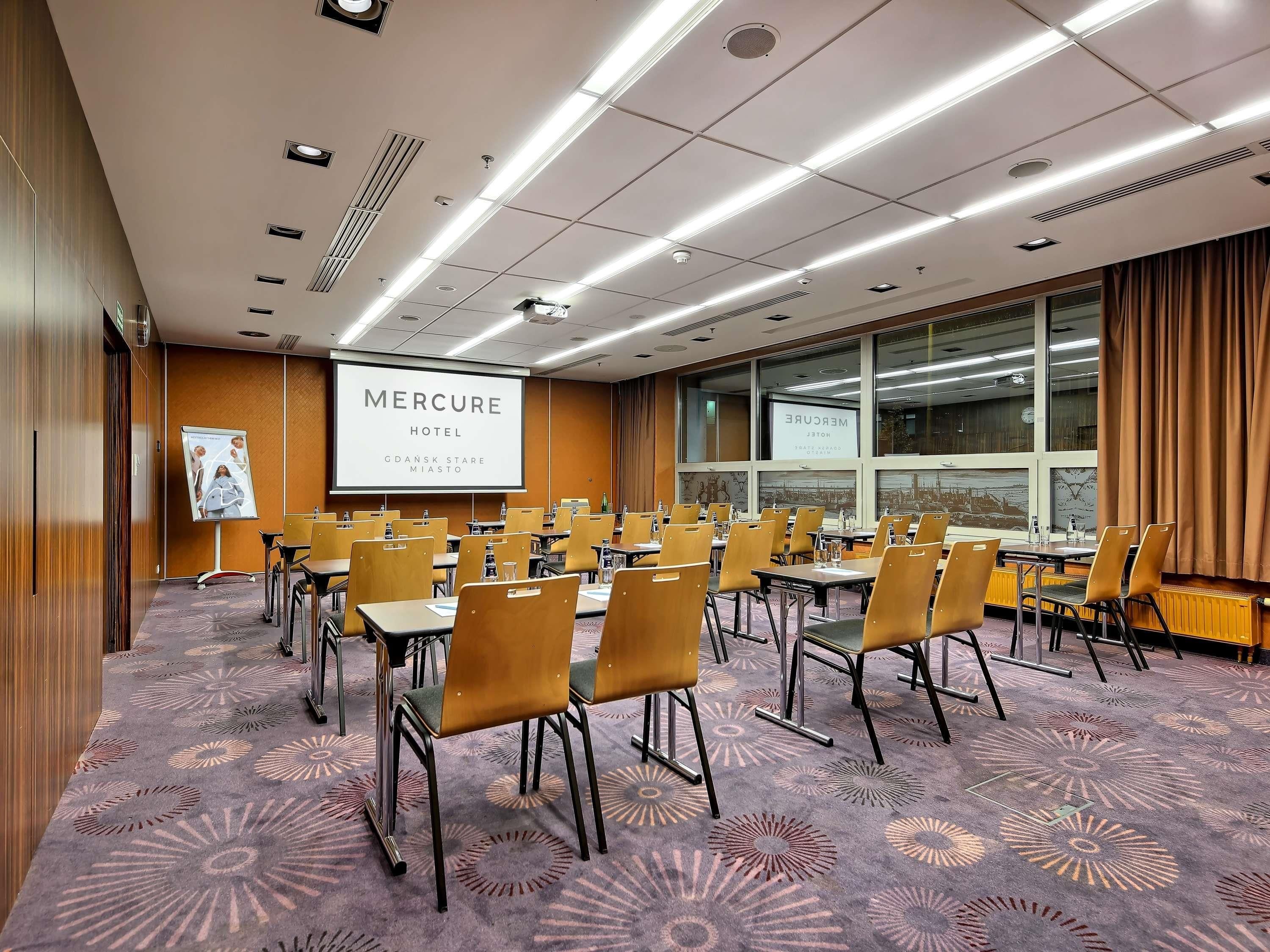 A conference room with a large screen displaying 'MERCURE HOTEL,' rows of chairs, and a patterned carpet.