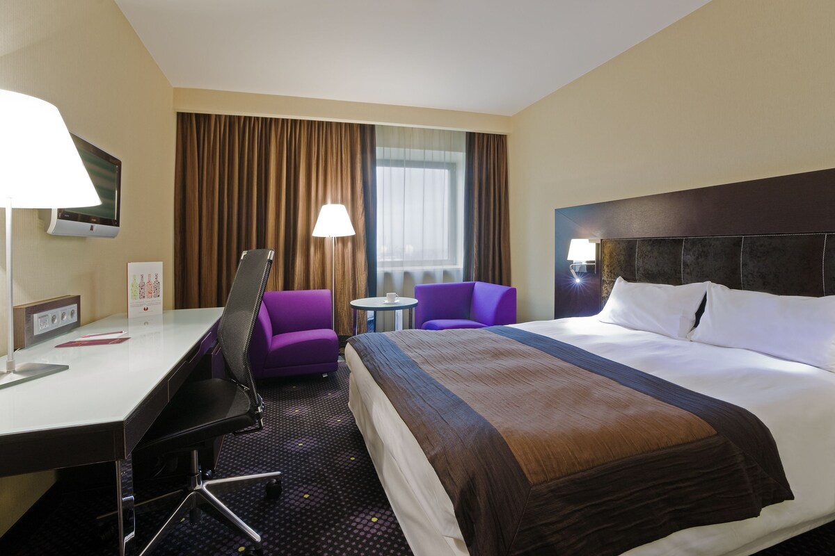 A hotel room with a large bed, a desk with a lamp, two purple chairs, a small table, and a TV mounted on the wall.