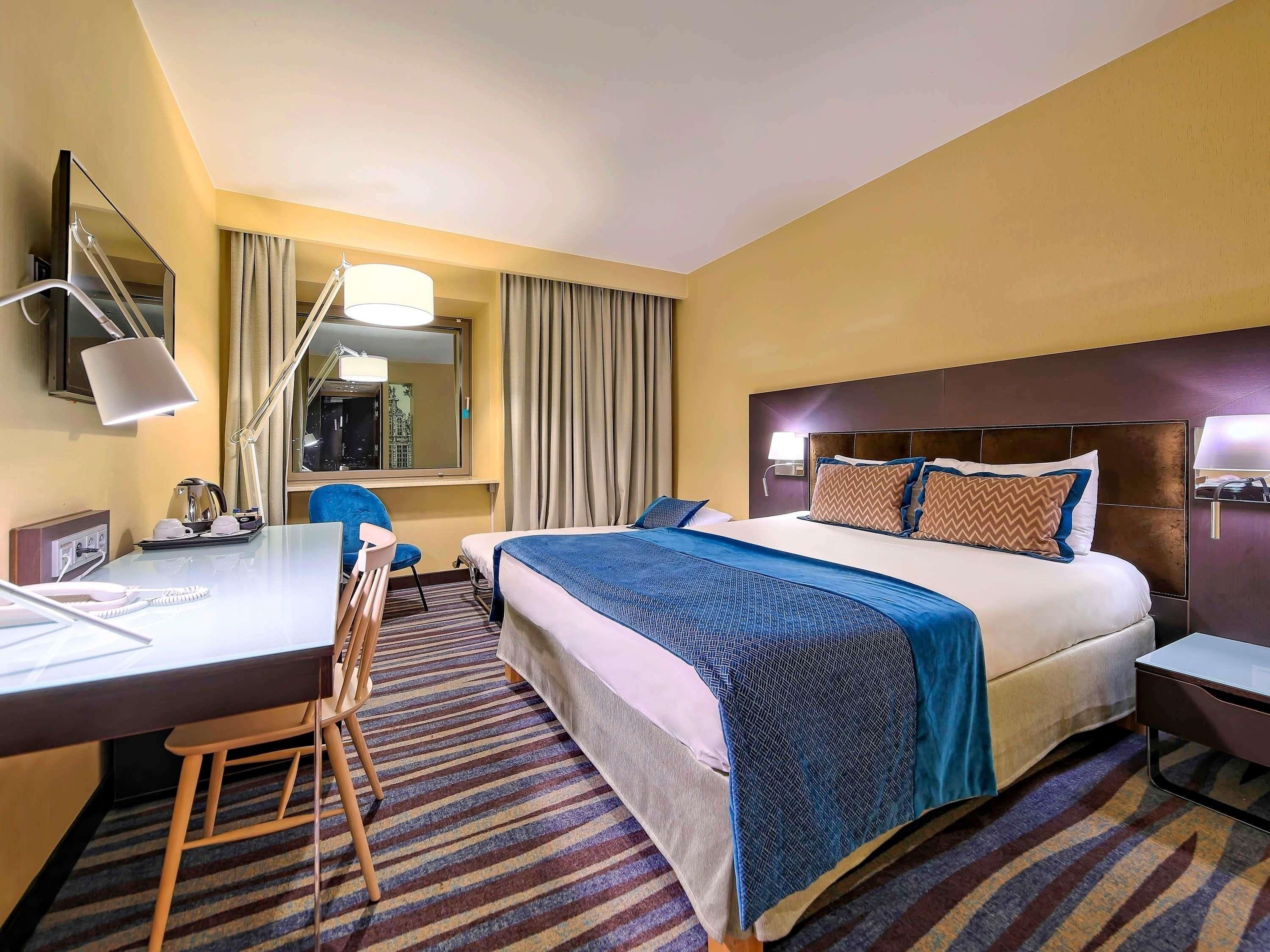 Habitación de hotel con cama, escritorio, silla y ventana con cortinas.