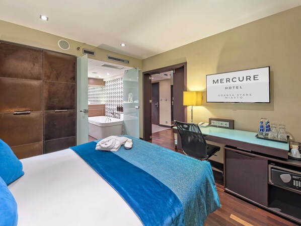 A hotel room with a bed, a desk, a chair, a minibar, and a bathroom visible through an open door.