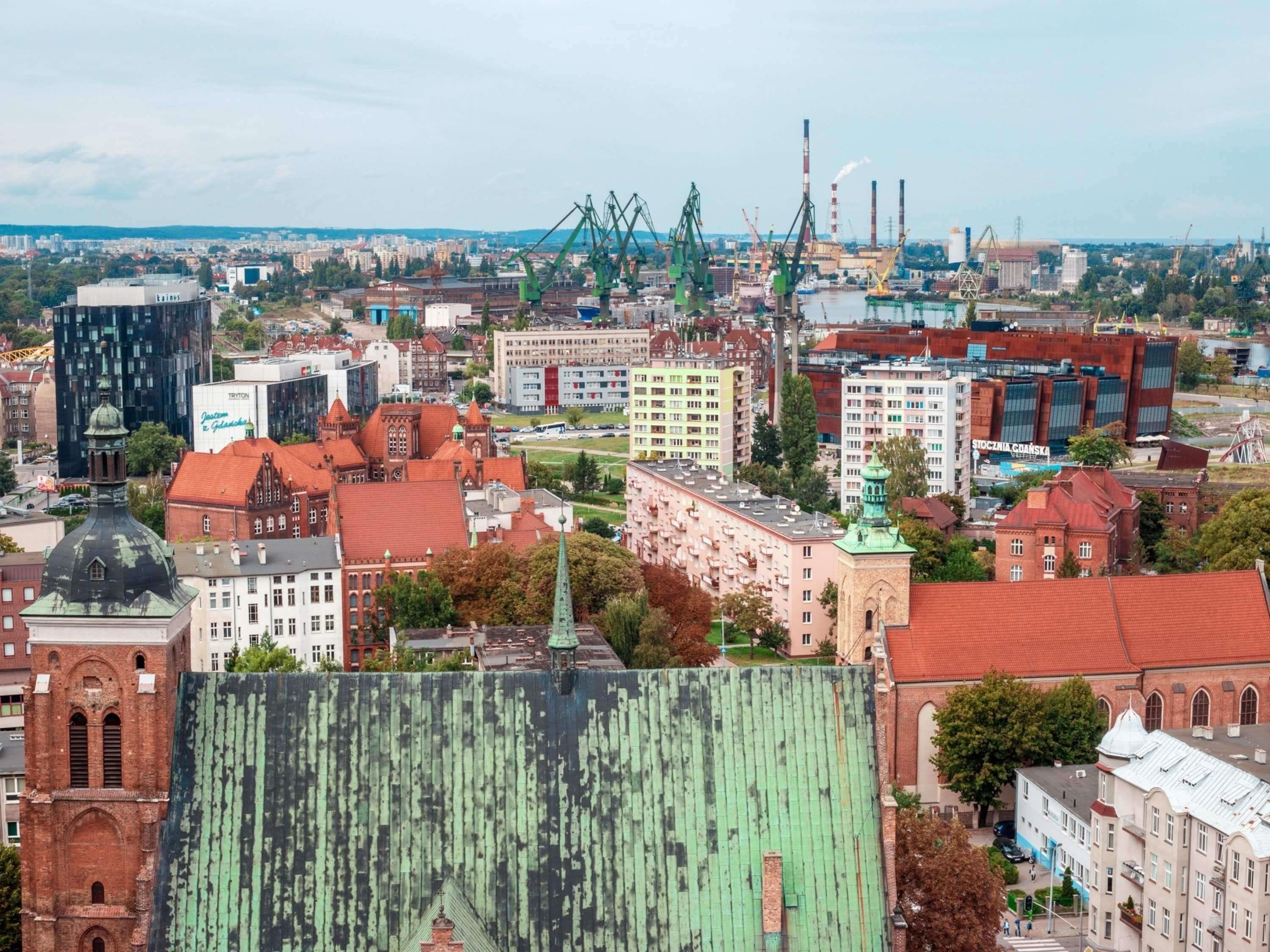 A cityscape with a mix of historical and modern buildings, including a prominent church with a green dome and a large industrial area with cranes and smokestacks.