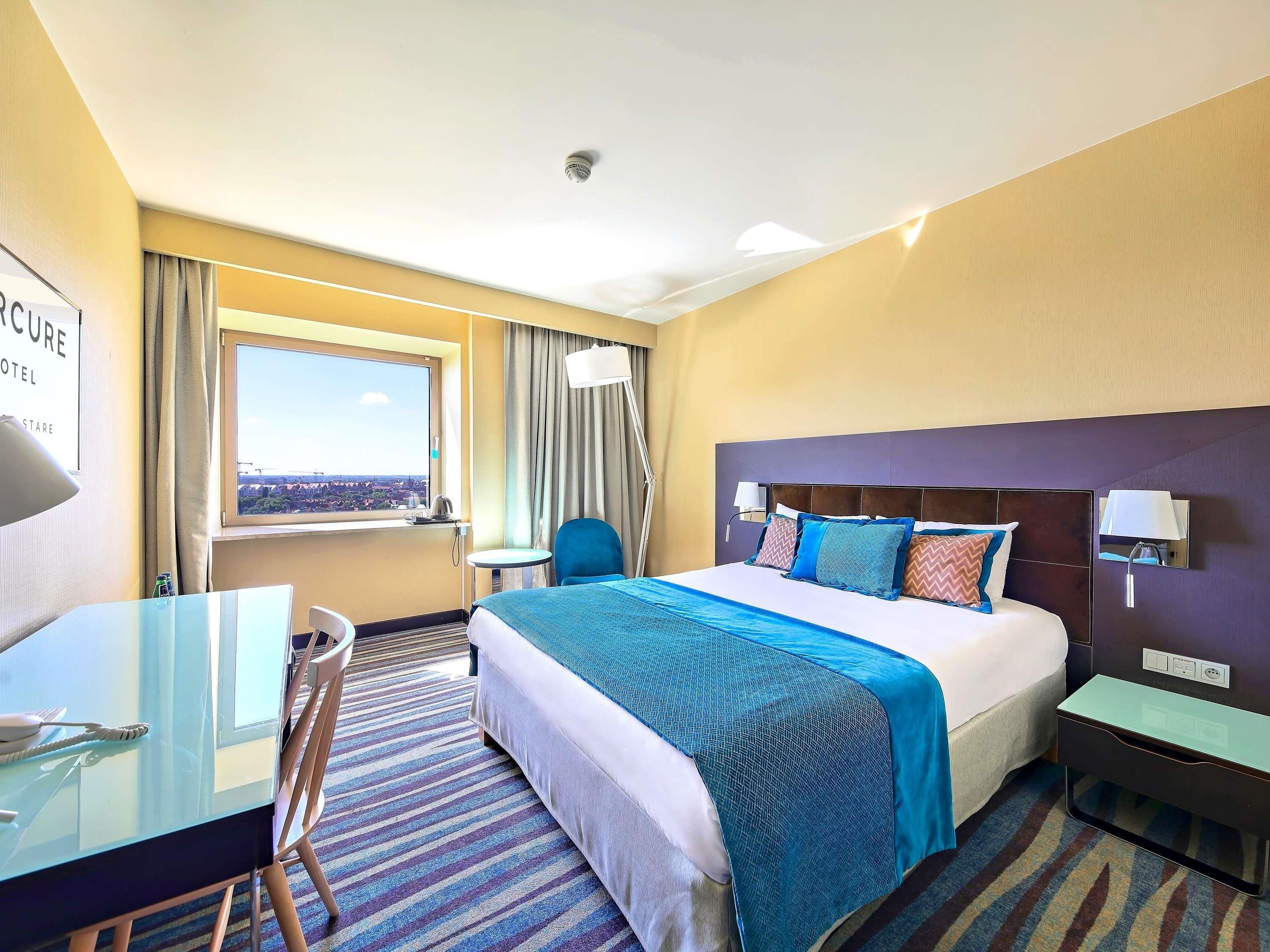 A hotel room with a bed, a desk, a chair, a window with a view, and a ceiling-mounted smoke detector.