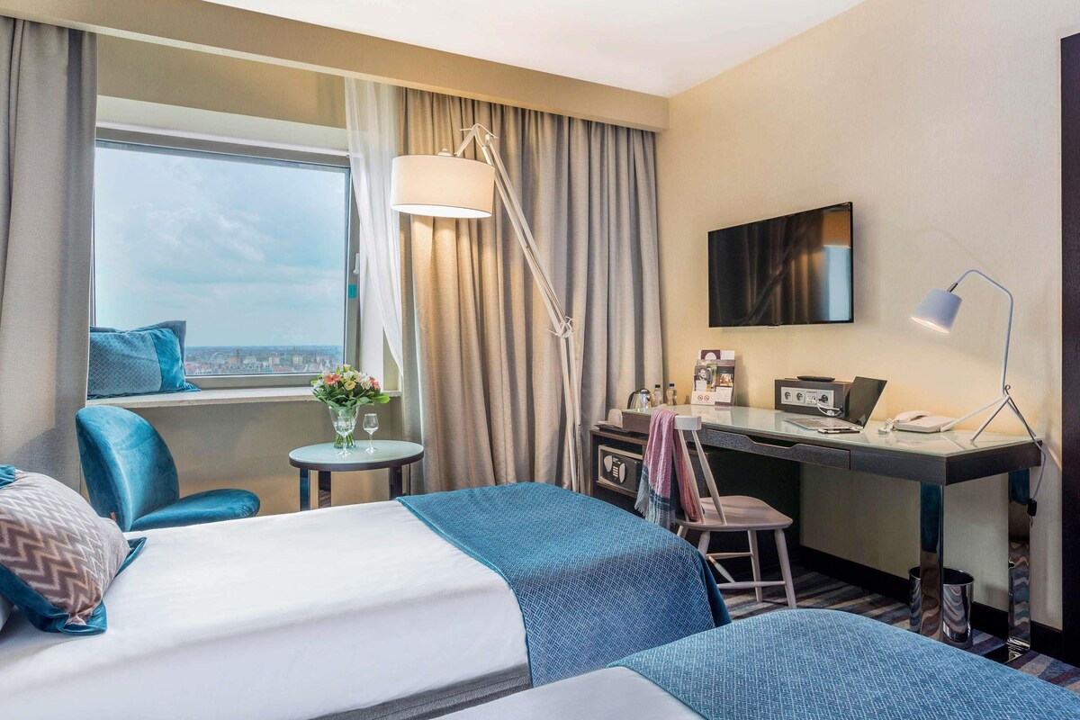 Une chambre d’hôtel avec deux lits, un bureau, une chaise, une télévision et une fenêtre donnant sur un beau paysage.