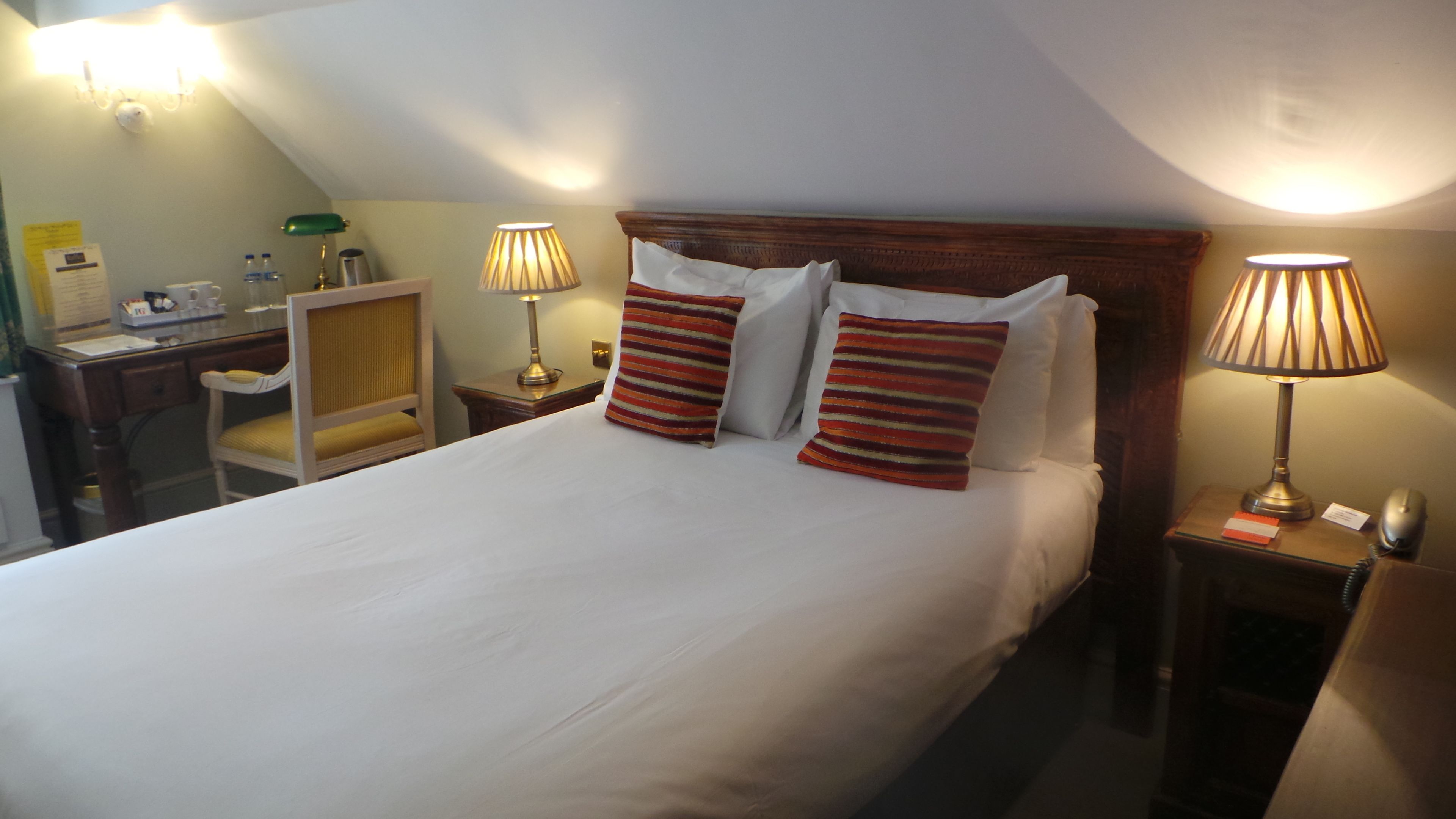 A hotel room with a large bed, two bedside lamps, a wooden headboard, a desk with a lamp, and a chair.