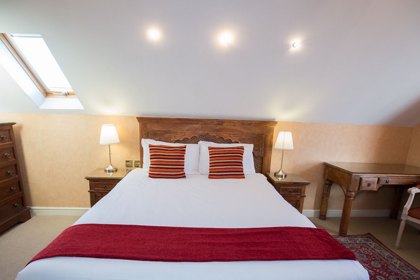 Un dormitorio ordenado con una cama grande, dos mesitas de noche con lámparas, un escritorio de madera y una manta roja.