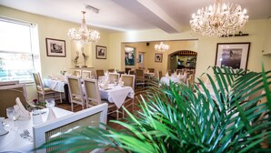 Une salle à manger avec plusieurs tables dressées pour les convives, des lustres, des tableaux encadrés aux murs et une grande plante en pot au premier plan.