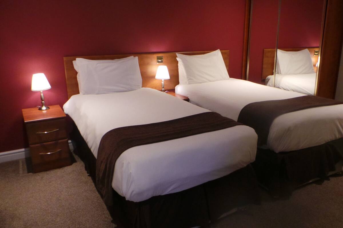 A hotel room with two beds, a nightstand with a lamp, and a radiator.
