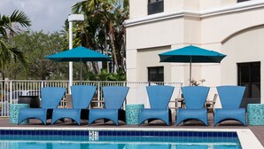 A poolside area with blue chairs and umbrellas, a tiled pool, and a white building with windows. - Hampton Inn & Suites by Hilton Miami-Doral/Dolphin Mall (Miami)