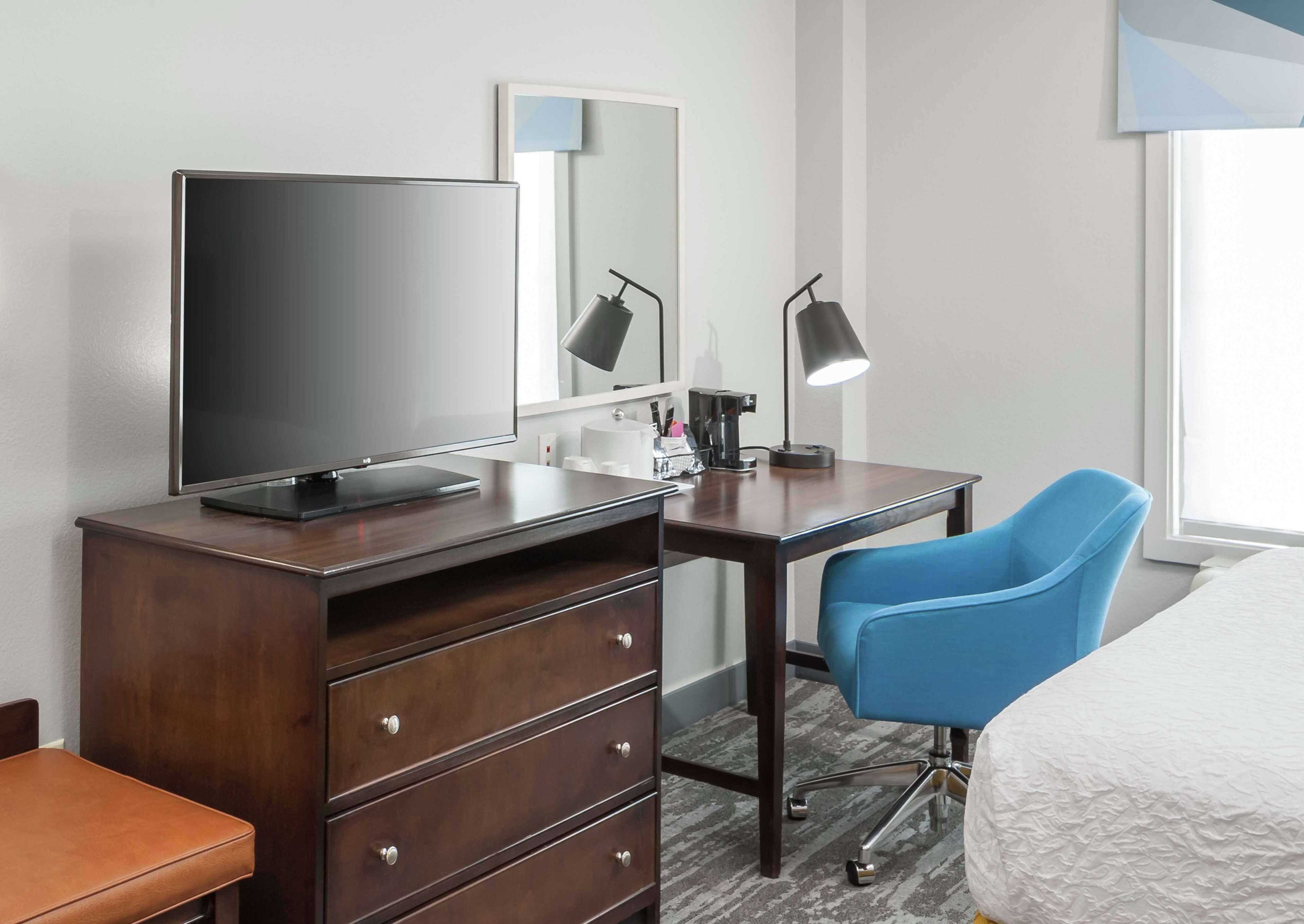 Hotellihuone, jossa on puinen työpöytä, televisio, sininen tuoli, sänky ja ikkuna.