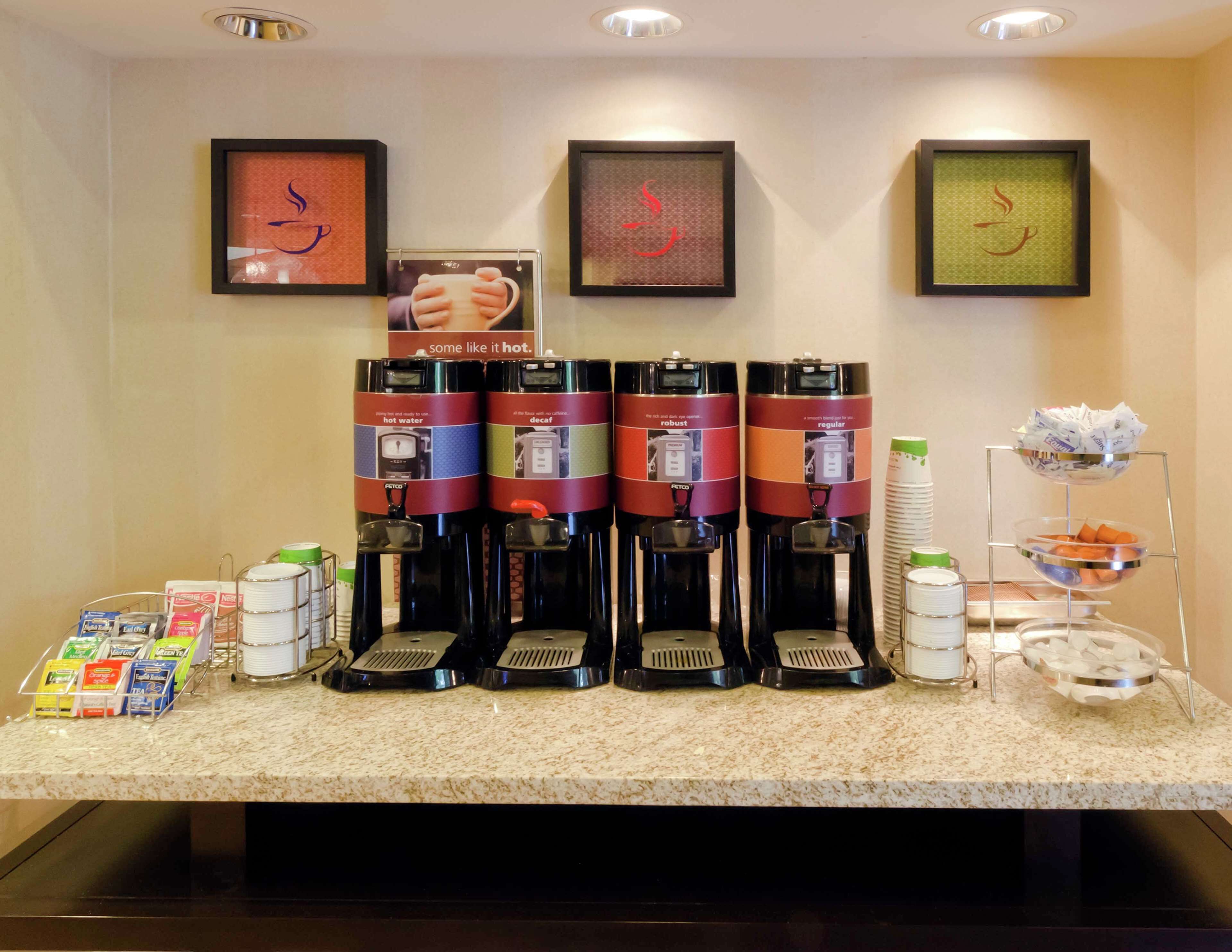 A coffee station with four coffee makers, a stack of disposable cups, and a tray with snacks.