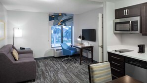 A modern hotel room with a gray sofa, a blue office chair, a desk with a lamp, a large window with a view of palm trees, a microwave, and a dishwasher. - Hampton Inn & Suites by Hilton Miami-Doral/Dolphin Mall (Miami)