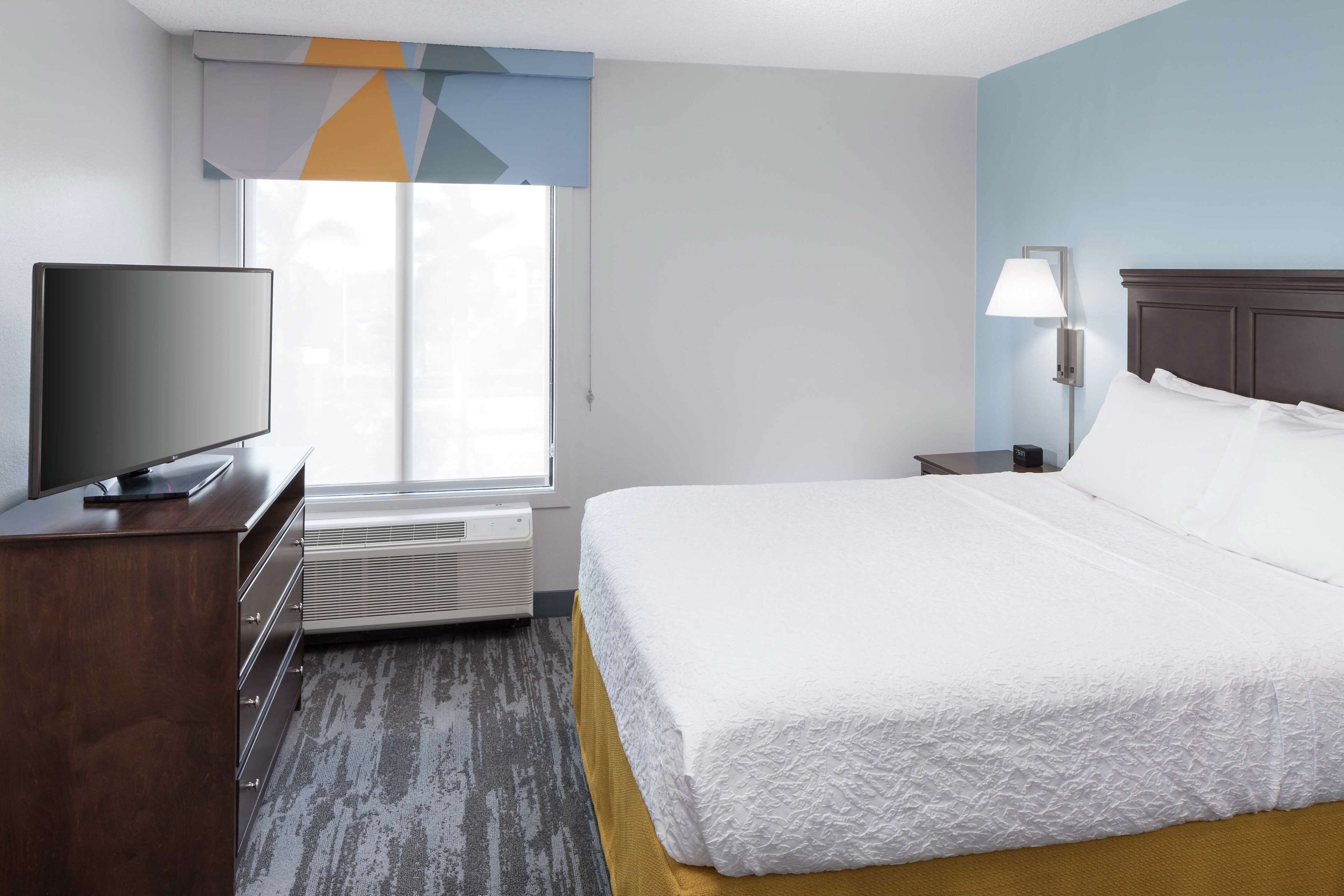 A hotel room with a bed, a television on a wooden dresser, a lamp, and a window with blinds.