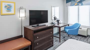 Habitación de hotel con televisión de pantalla plana, un escritorio con lámpara, una silla azul, un banco y una cama.