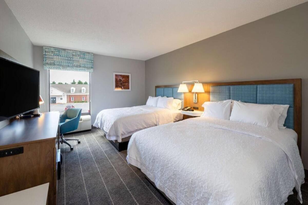 A hotel room with two beds, a desk, a television, and a window with a view of buildings.