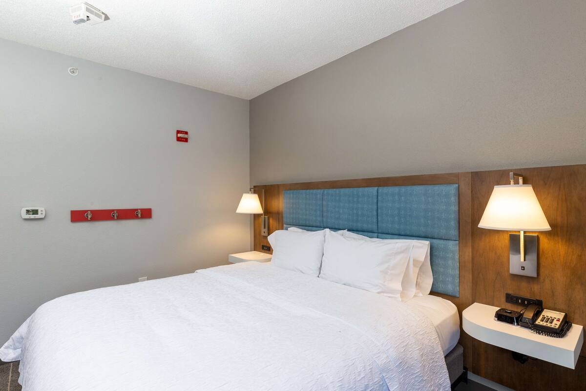 A hotel room with a large bed, two bedside lamps, a nightstand, a red emergency exit sign, and a thermostat on the wall.