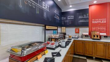 A breakfast buffet area with a variety of food items, including a hot food station, and a sign promoting waffles.