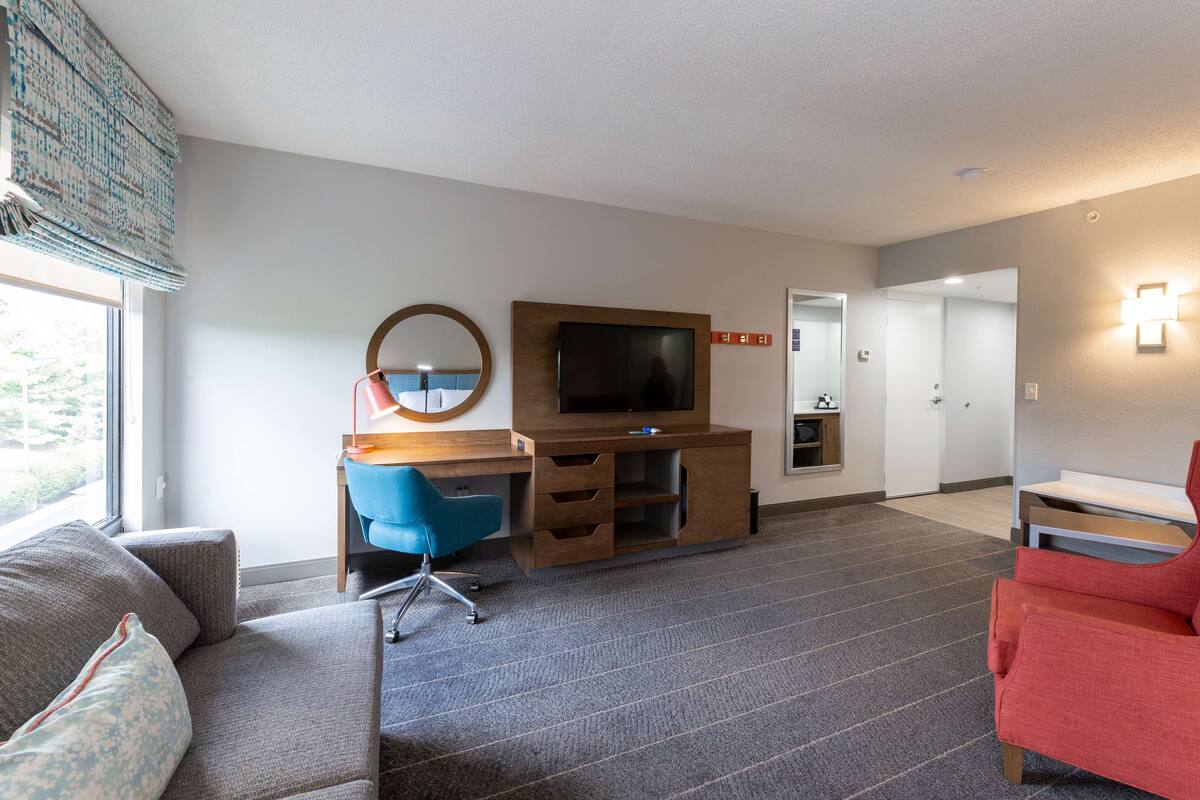A modern hotel room with a large flat-screen TV, a wooden entertainment unit, a blue office chair, a grey sofa, and a red armchair.