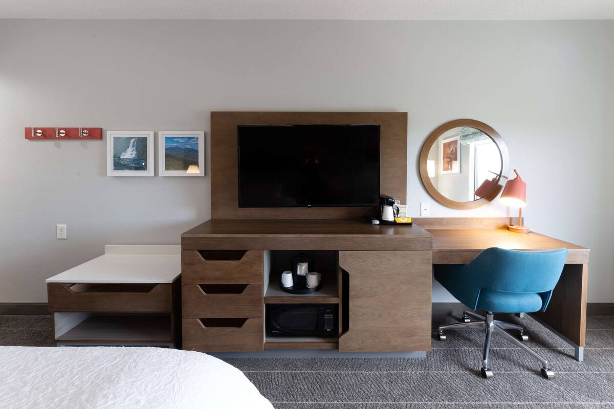 A hotel room with a wooden desk, a flat-screen TV, a mirror, a blue chair, a bed, and a microwave.