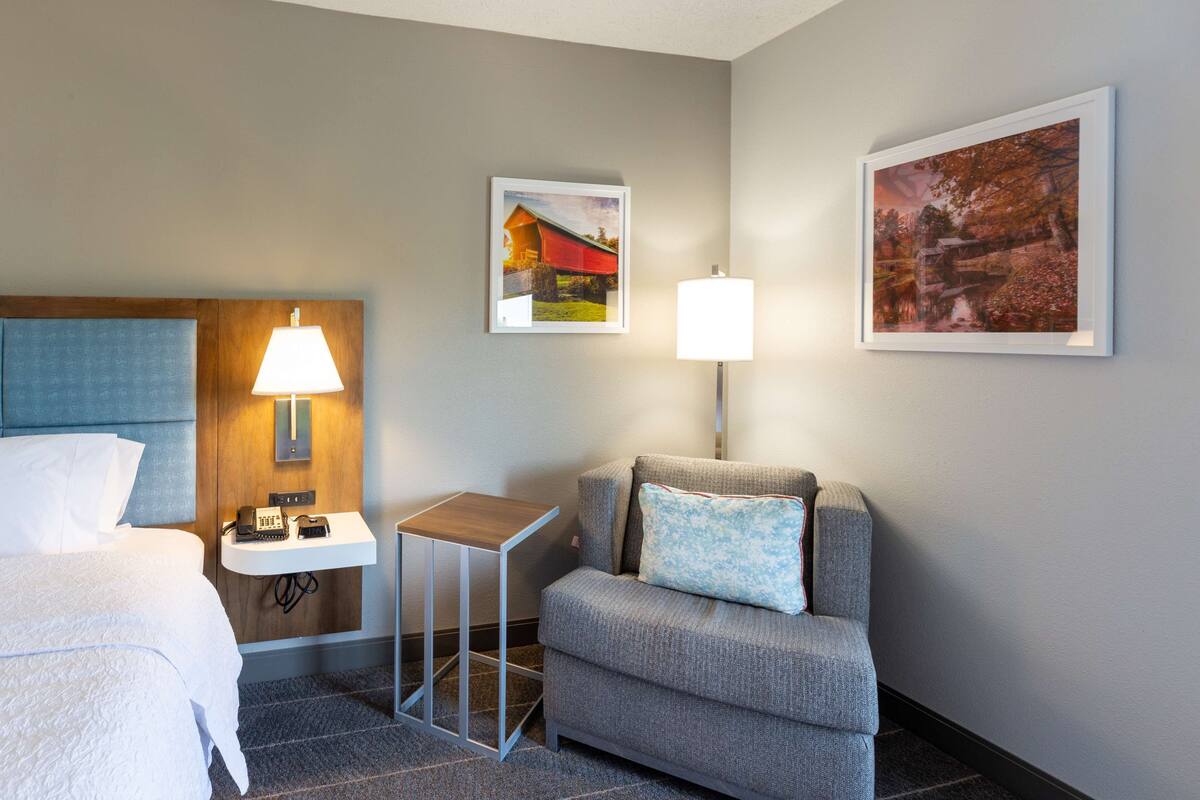 A hotel room with a bed, a desk with a lamp, a small table, a sofa with a pillow, and two framed pictures on the wall.