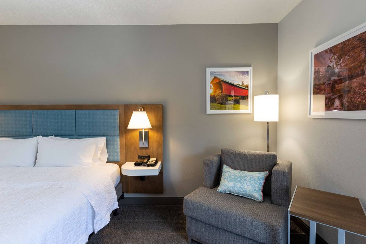 A hotel room with a bed, a desk, a chair, a nightstand with a lamp, and two framed pictures on the wall.