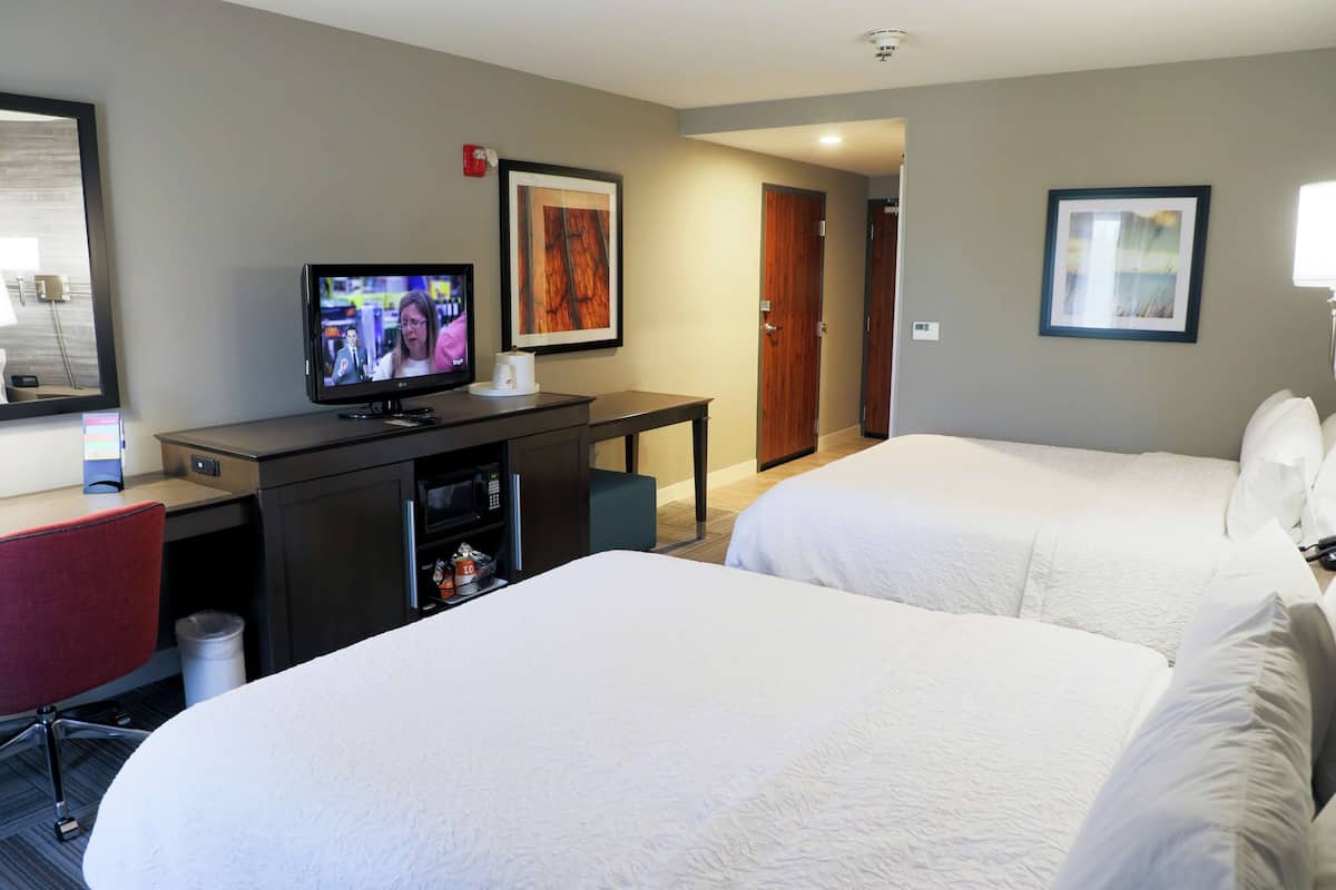 A hotel room with two beds, a desk with a television, a chair, and a small trash bin.