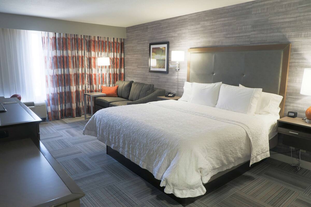 A hotel room with a large bed, a desk, a chair, a sofa, and a nightstand with a lamp.