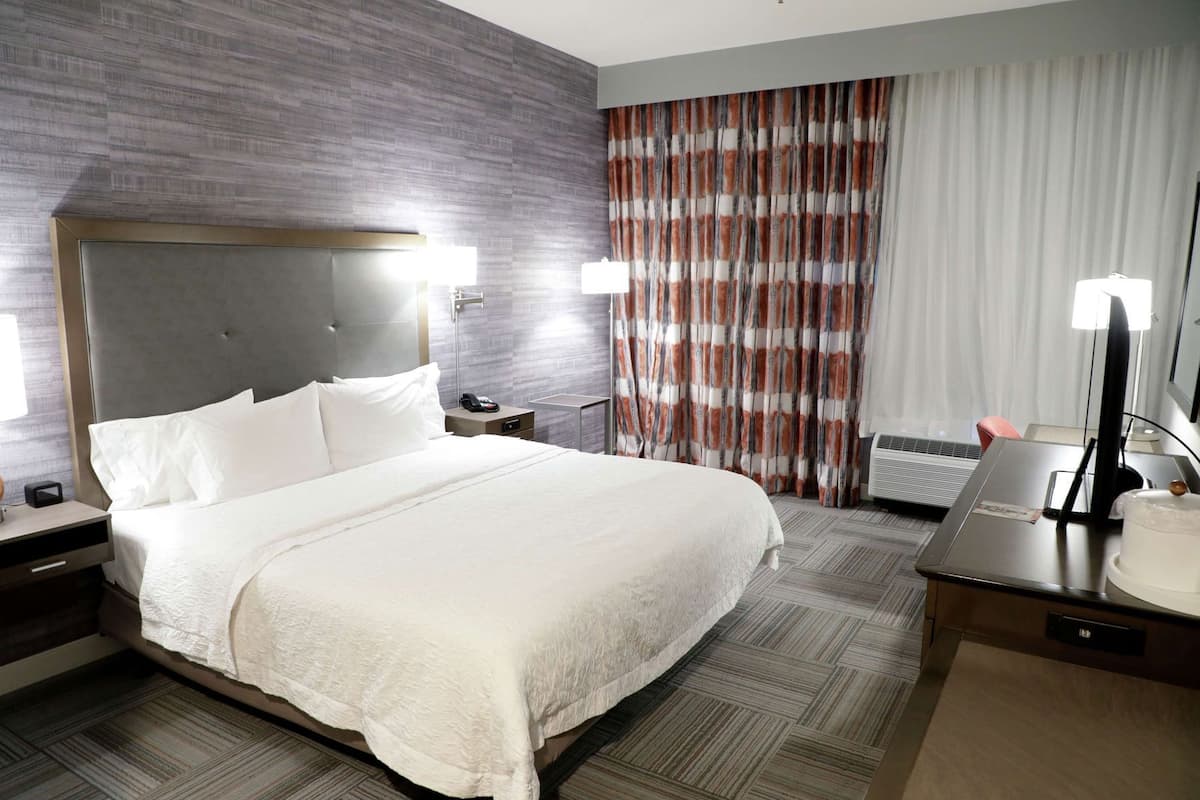 A hotel room with a large bed, a nightstand with a lamp, a desk with a computer, and a window with curtains.