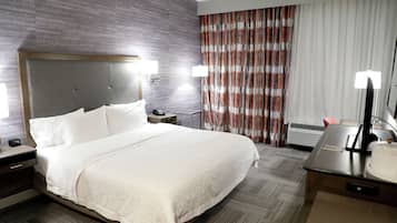 A hotel room with a large bed, a desk with a lamp, a nightstand, and a window with curtains.