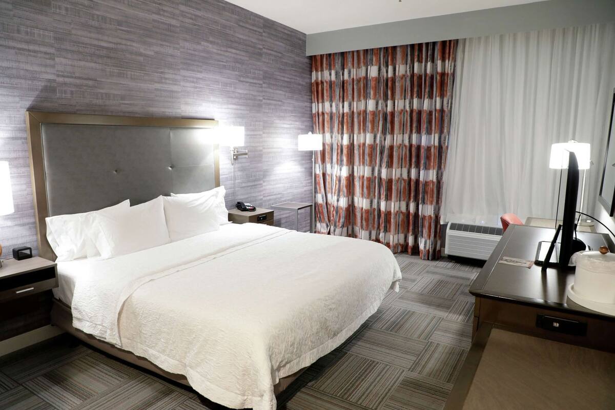 A hotel room with a large bed, a nightstand with a lamp, a desk with a computer, and a window with curtains.