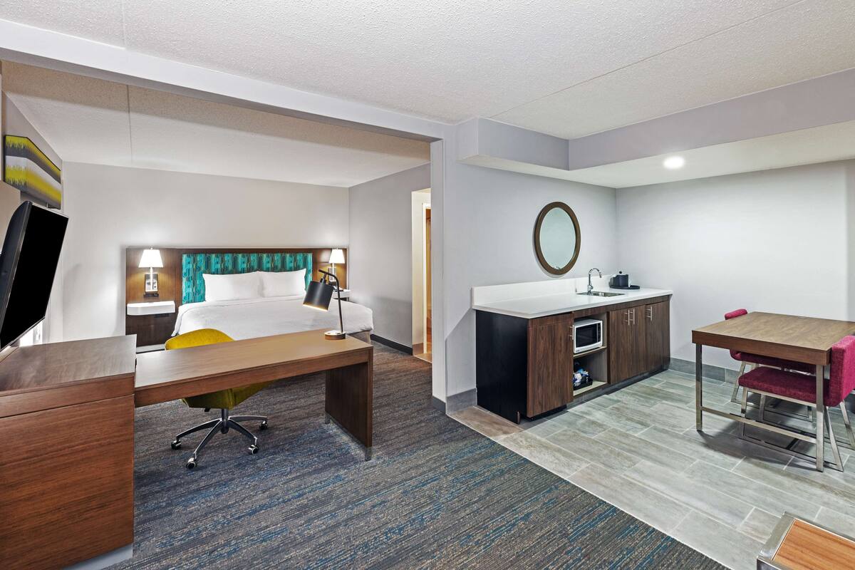 A hotel room with a bed, desk, chair, kitchenette, and a dining table.