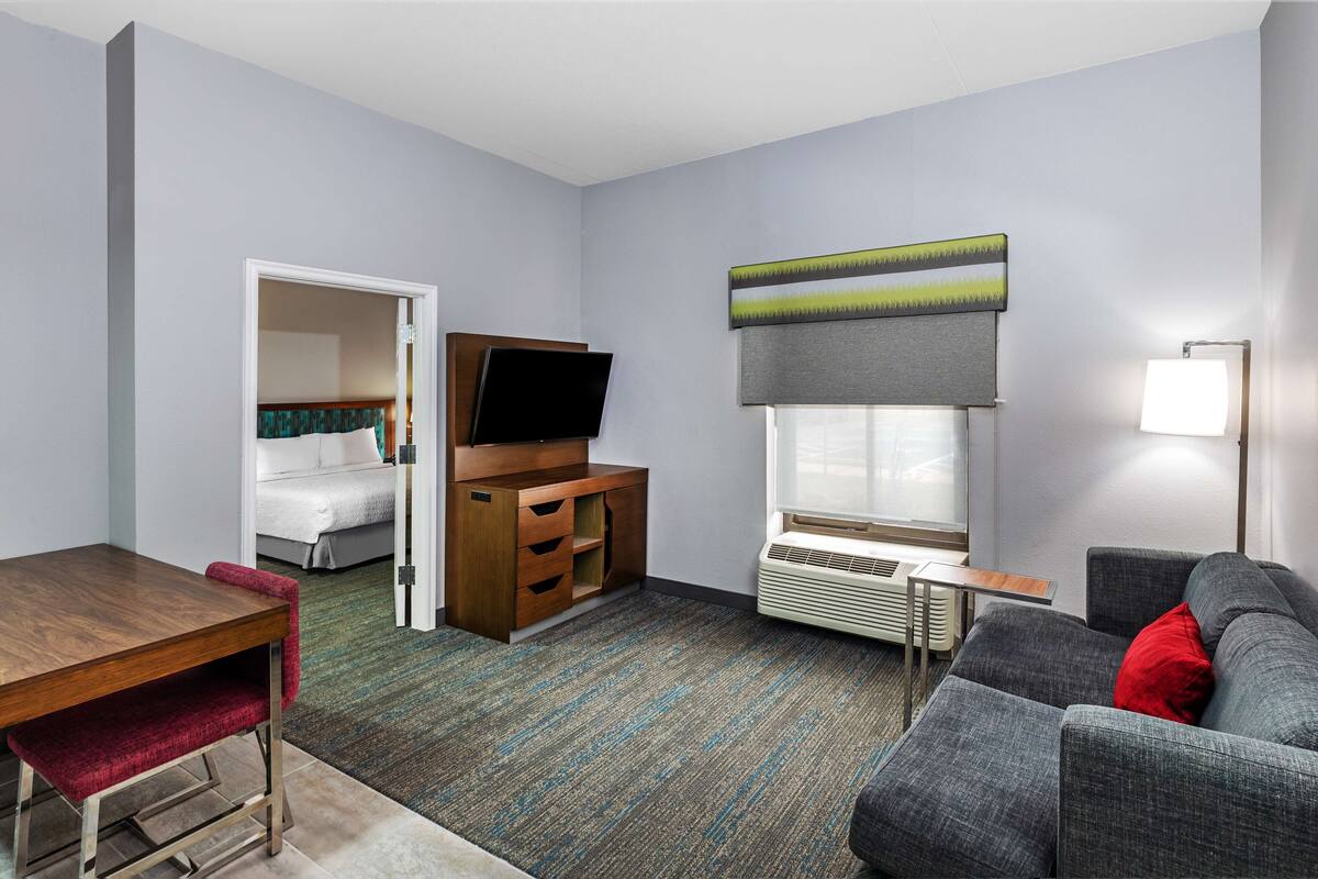 A hotel room with a bed, a TV, a desk, a chair, a sofa, and a window with blinds.