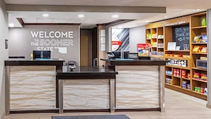 Zona reception di un hotel con un cartello 'Welcome to the Sooners', una postazione self-service e scaffali con snack.