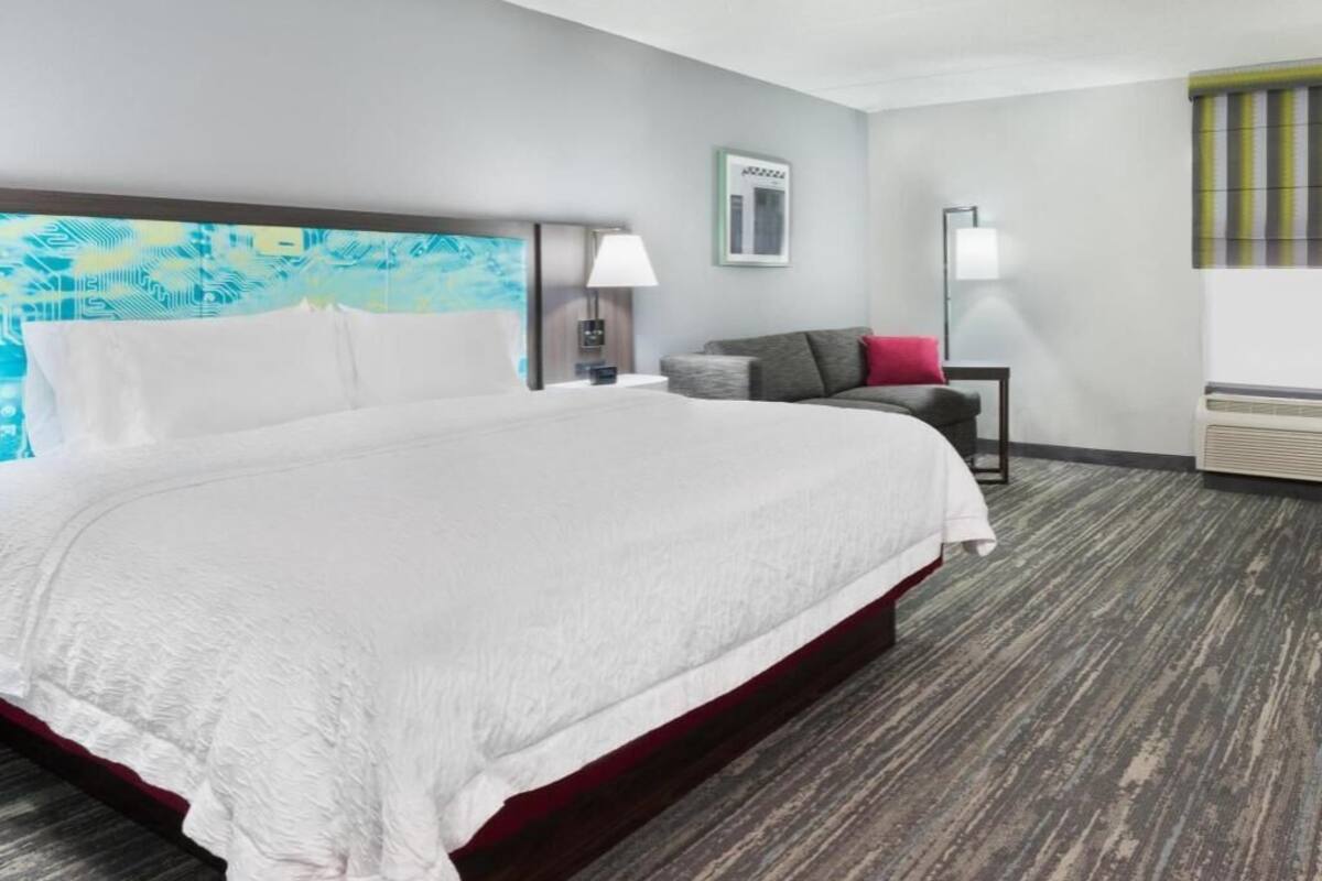 A hotel room with a large bed, a nightstand, a lamp, a small table, and a window with blinds.