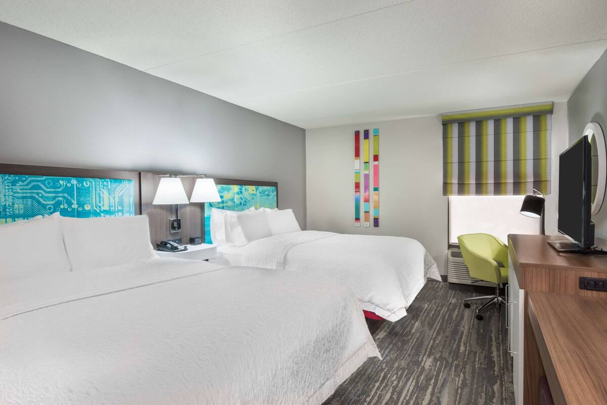 A hotel room with two beds, a desk, a chair, a TV, and a window with blinds.