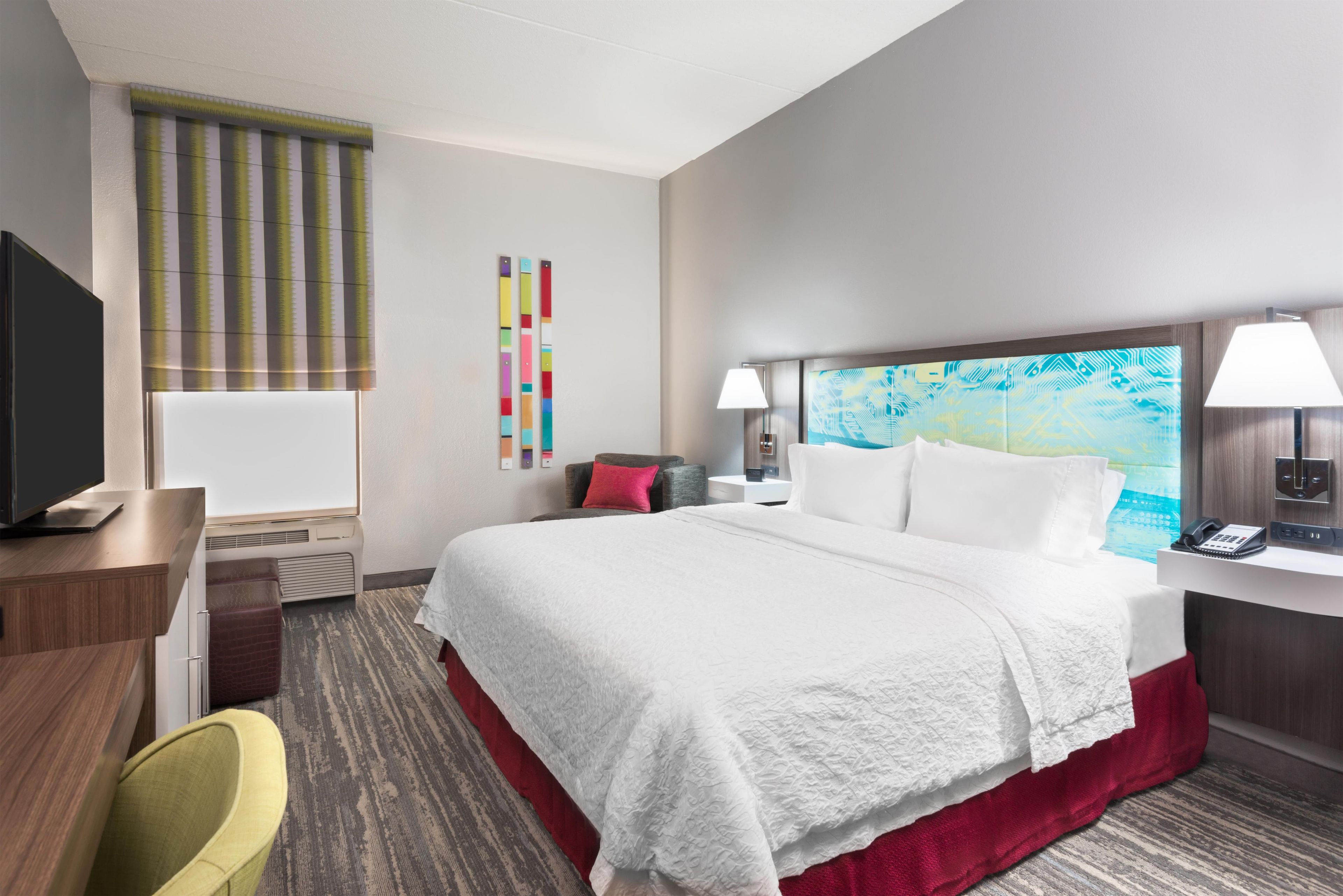 Habitación de hotel con cama, escritorio, silla, televisor y un cuadro en la pared.