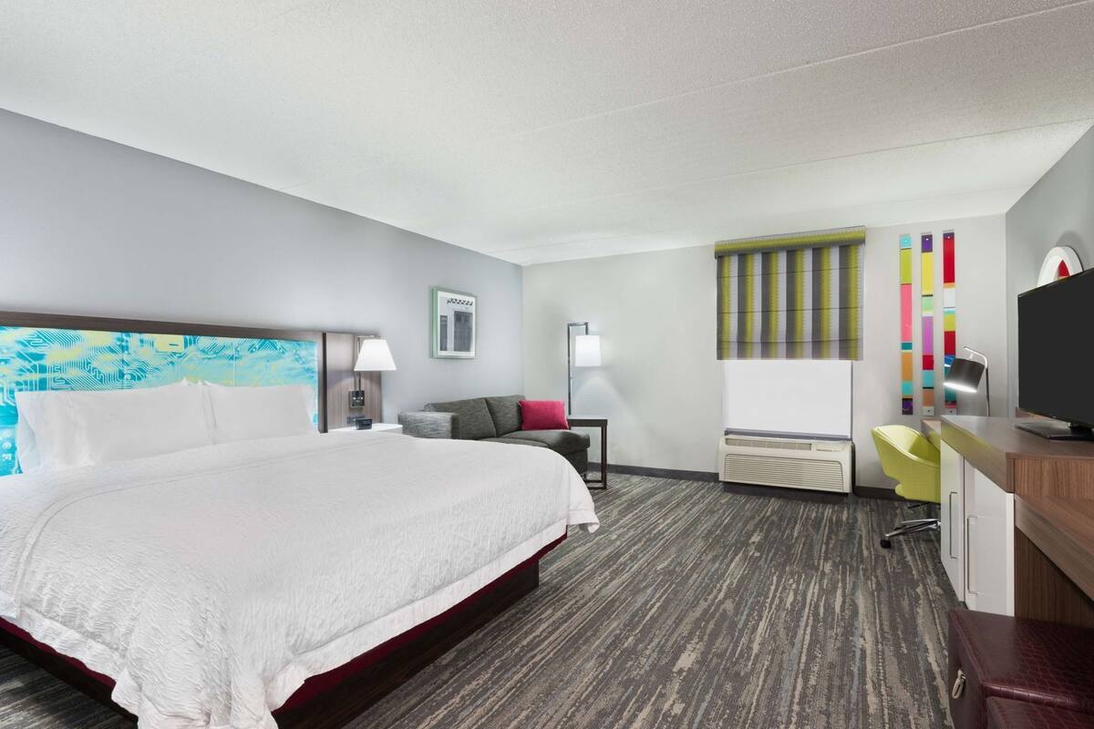 A hotel room with a large bed, a desk with a chair, a TV, and a window with blinds.