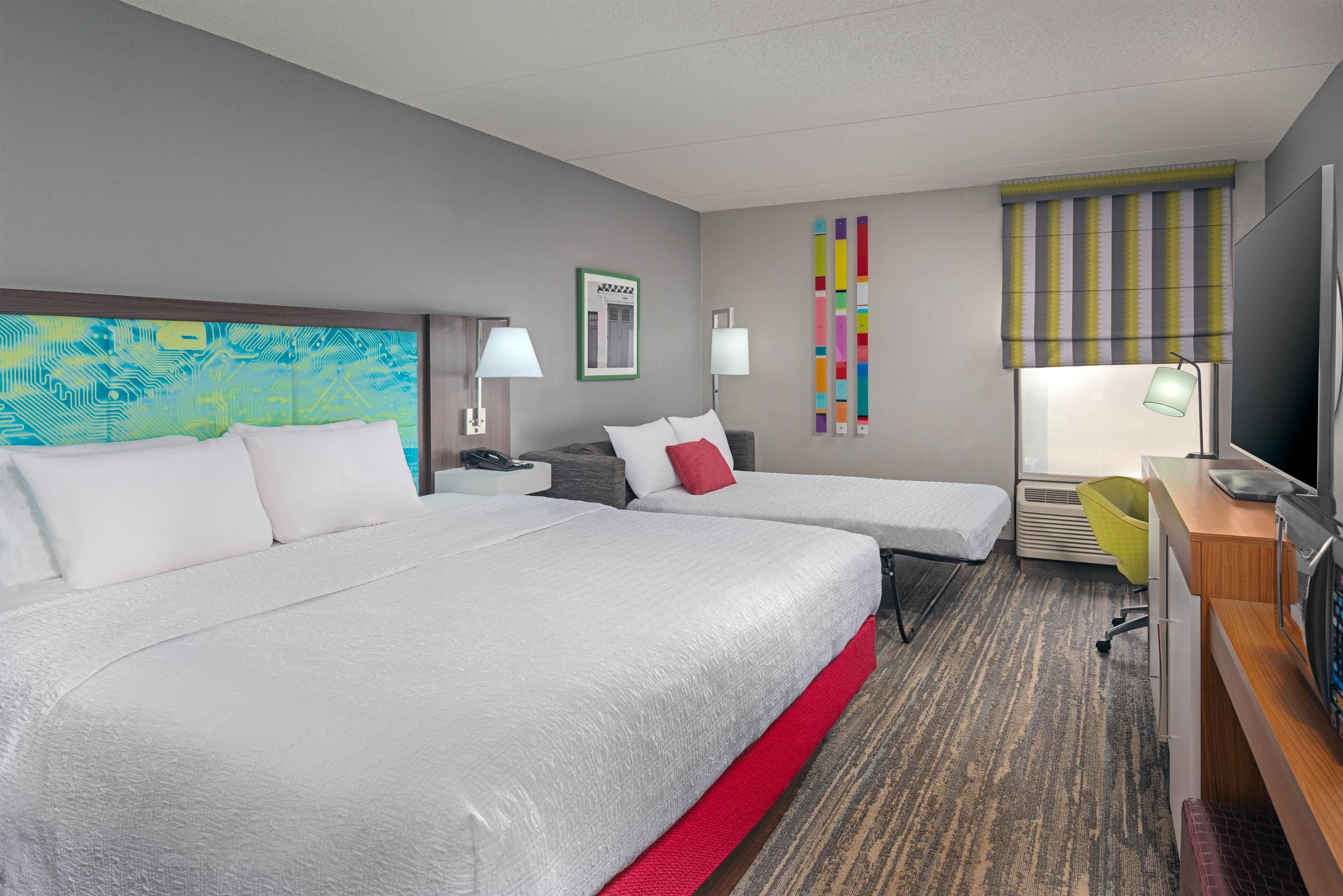 A hotel room with two beds, a desk, a chair, a TV, and a window with blinds.
