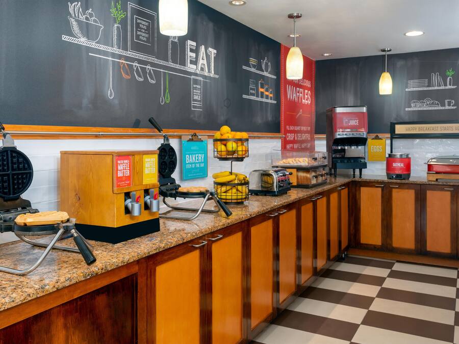 A breakfast bar with waffle makers, a juice dispenser, and a sign indicating a bakery item of the day.