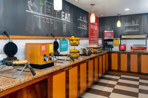 A breakfast bar with waffle makers, a juice dispenser, and a sign indicating a bakery item of the day. - Hampton Inn Ellenton/Bradenton (Ellenton)