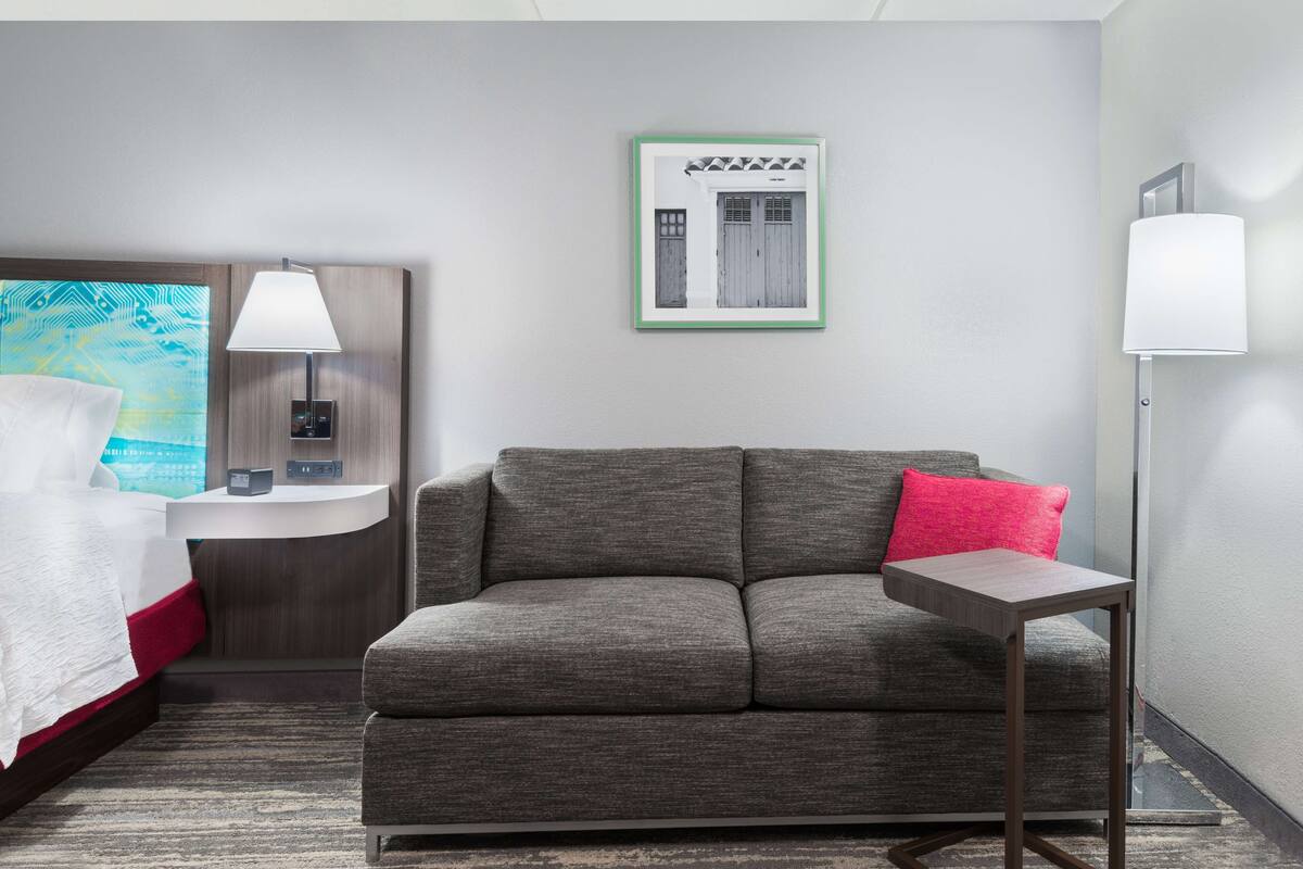 A hotel room with a grey sofa, a small table, a bedside table with a lamp, a framed picture on the wall, and a bed with a red accent.
