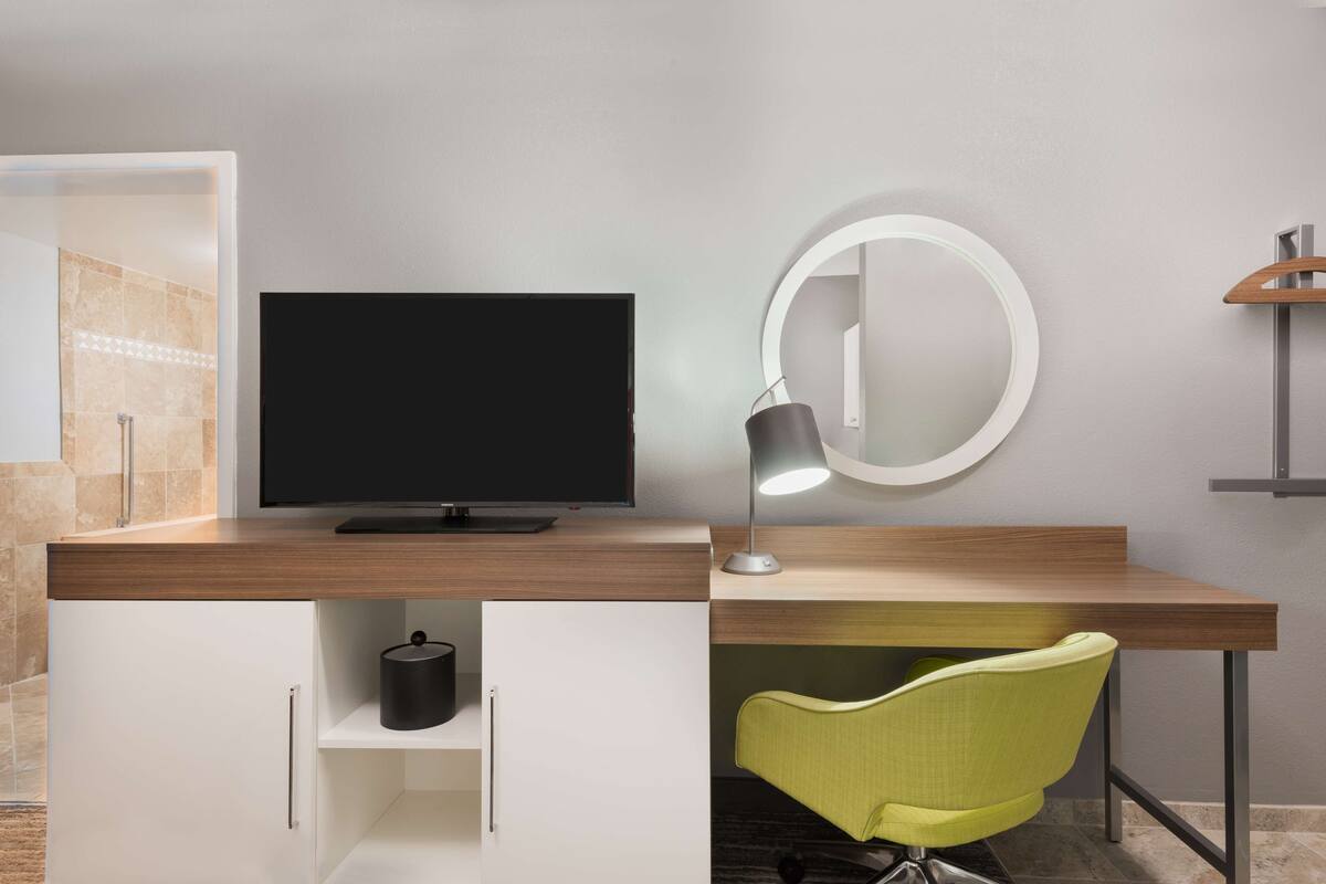 A modern hotel room with a wooden desk, a flat-screen TV, a desk lamp, a mirror, and a green chair.