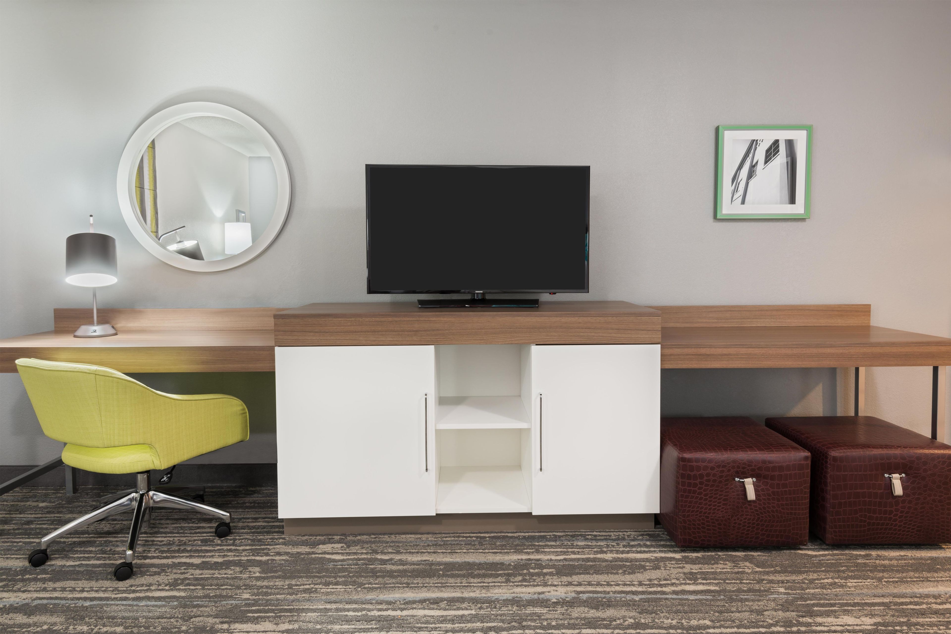 Habitación de hotel con escritorio de madera, televisor, espejo, una silla amarilla y un cuadro en la pared.