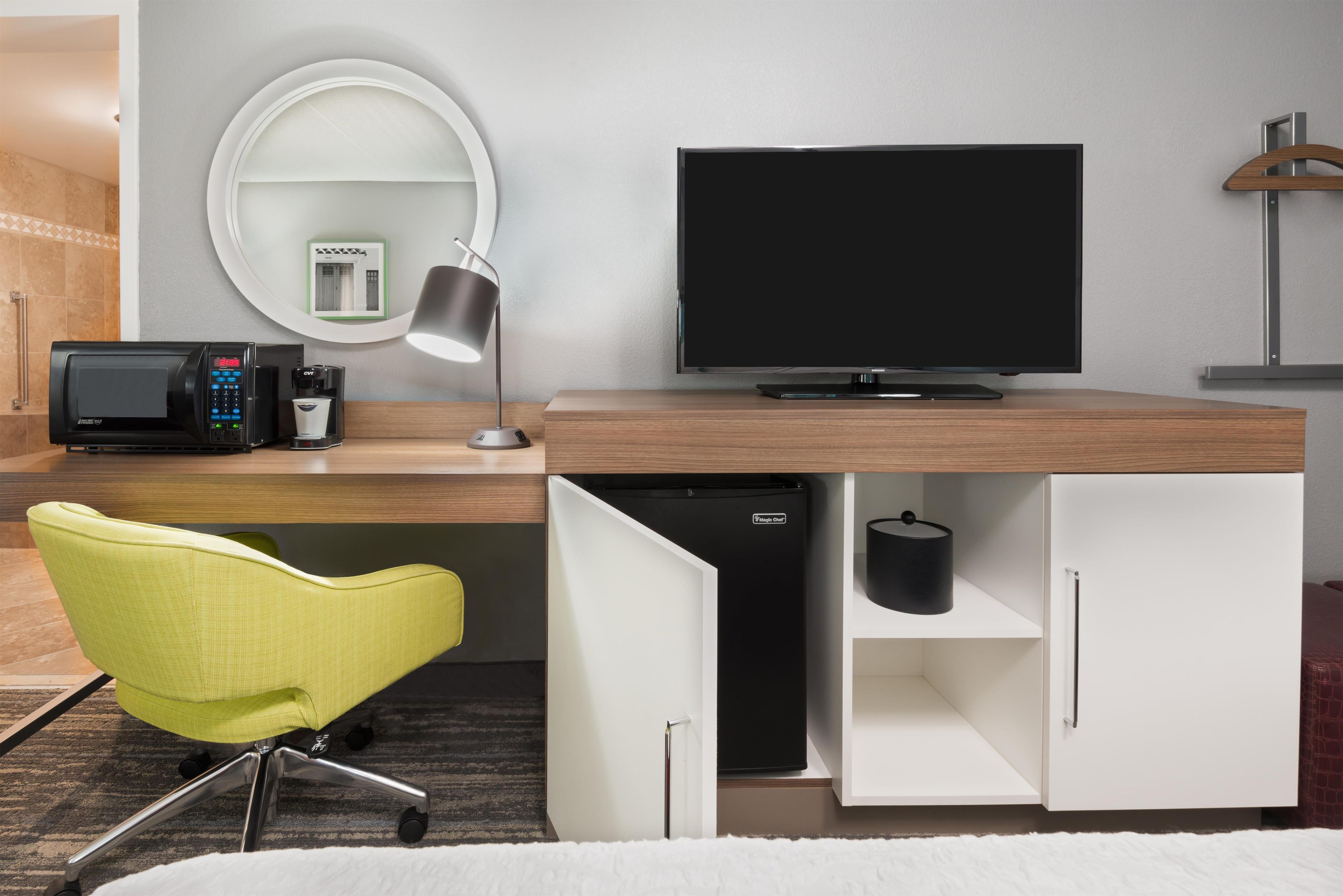 Habitación de hotel moderna con televisión de pantalla plana, microondas, cafetera, escritorio con lámpara, silla y armario.