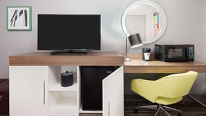 A hotel room with a wooden desk, a flat-screen TV, a microwave, a coffee maker, a mini-fridge, a yellow chair, and a mirror with a light. - Hampton Inn Ellenton/Bradenton (Ellenton)