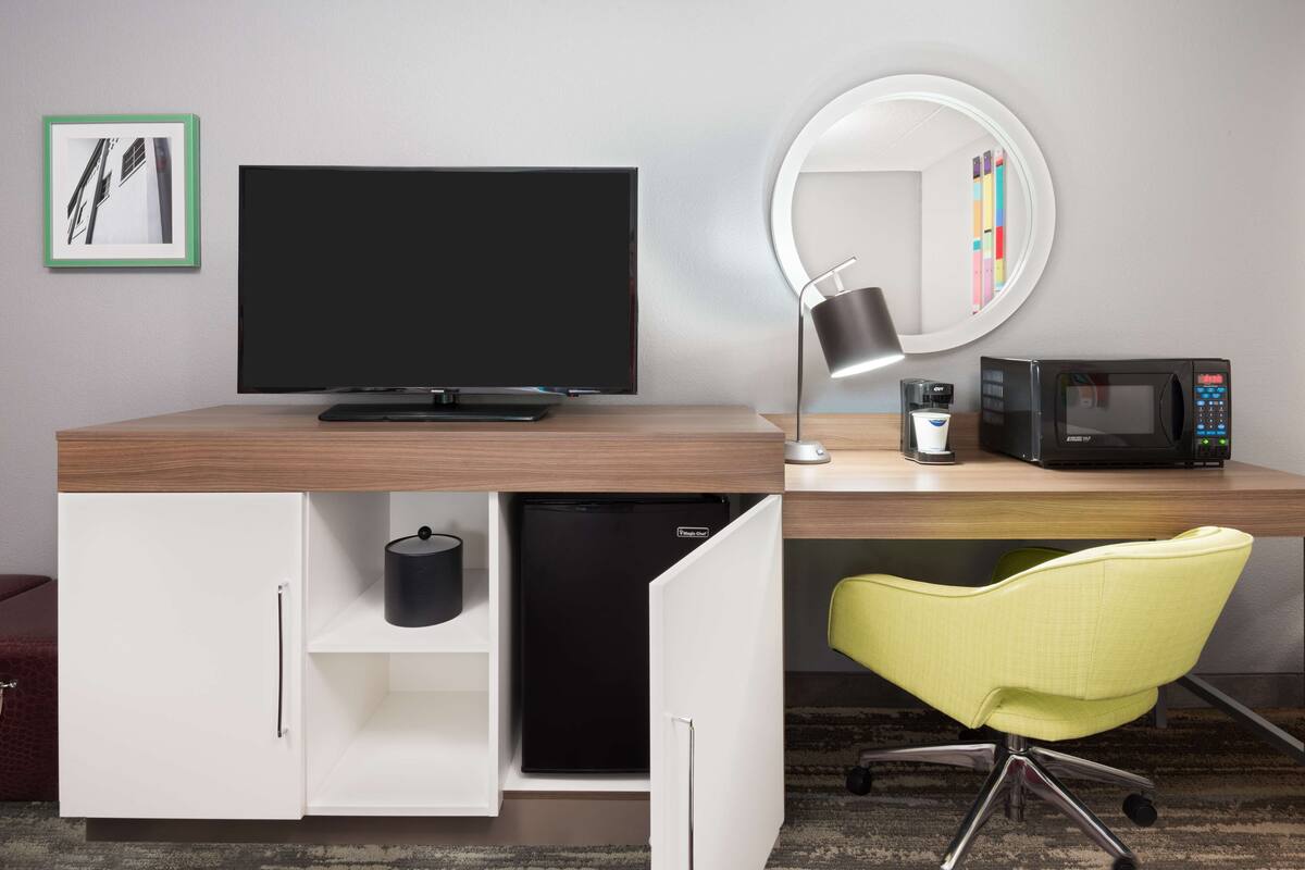 A hotel room with a wooden desk, a flat-screen TV, a microwave, a coffee maker, a mini-fridge, a yellow chair, and a mirror with a light.
