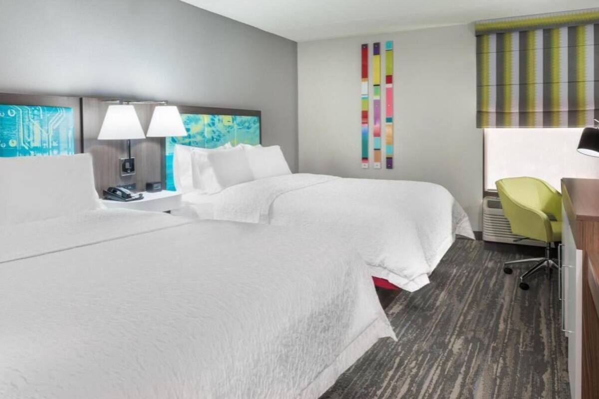 A hotel room with a large bed, two bedside lamps, a desk with a chair, and a colorful wall decoration.