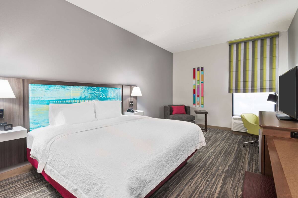 A hotel room with a large bed, a desk with a chair, a TV, a lamp, and a window with blinds.